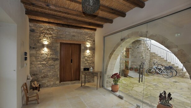 A stone-walled entrance with a wooden door, a small table with a lamp, and a chair. There are potted plants and a bicycle outside. - Pentasino Heritage Suites (Agios Theodoros)