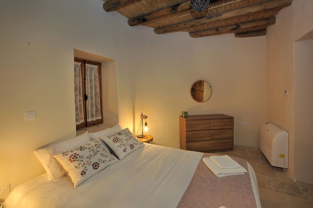A bedroom with a bed, a wooden dresser, a nightstand with a lamp, a window with curtains, and a ceiling with exposed beams.