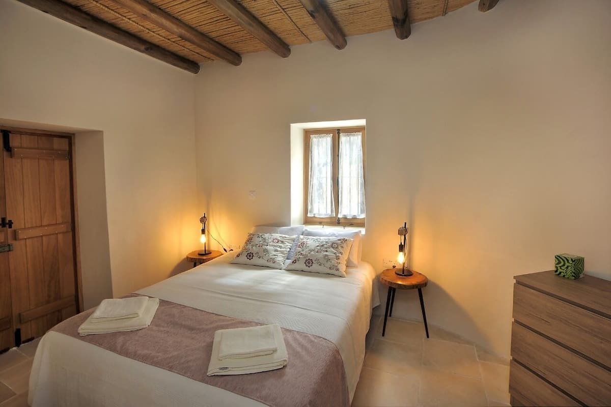 Un dormitorio con una cama, dos mesitas de noche con lámparas, un ventanal con cortinas, un armario de madera y un aparador.