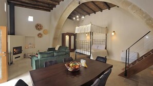A cozy living room with a fireplace, a dining table with a fruit bowl, a staircase, and a bed with a canopy. - Pentasino Heritage Suites (Agios Theodoros)