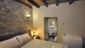 A bedroom with a stone wall, a bed with white linens, a nightstand with a lamp, and a door leading to a bathroom. - Pentasino Heritage Suites (Agios Theodoros)