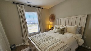 Une chambre à coucher avec un lit, des rideaux, une table de chevet, une lampe et une fenêtre.