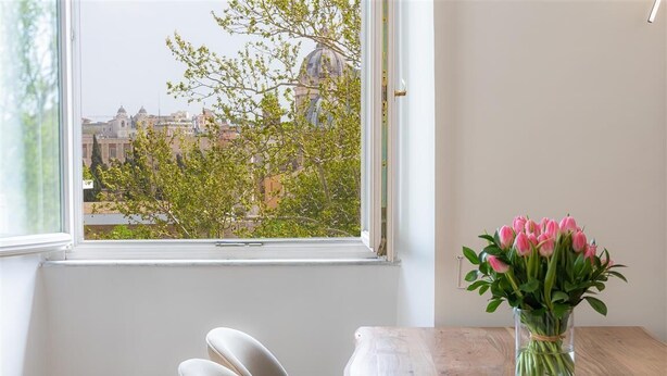 A room with a view of a cityscape through a window, a wooden table, and a vase of pink flowers.