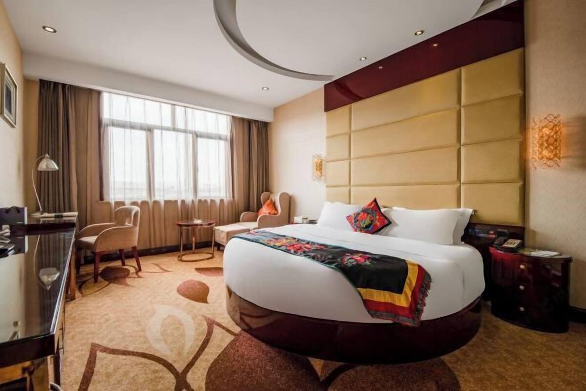A hotel room with a large bed, a desk, a chair, and a view of the city through large windows.