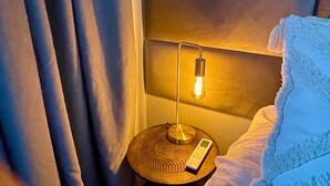 Un lit bien fait, avec un drap blanc et un oreiller, une table de chevet avec une lampe et une télécommande, et un rideau tiré à moitié sur le côté.