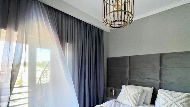 Une chambre moderne avec un grand lit, des rideaux gris et un luminaire suspendu original.