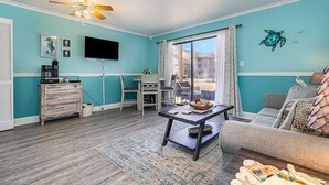 A living room with teal walls, a ceiling fan, a flat-screen TV, a dining table, and a sofa. - Cozy 1-bedroom condo with AC, WiFi in charming Little River. 3 miles from beach. (Little River)