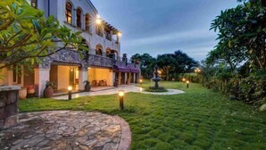 Un grand bâtiment élégant, doté d’une façade en pierre, de nombreux balcons et d’un jardin bien entretenu avec une fontaine.