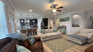 A spacious living room with a brown leather couch, a beige sofa, and a patterned rug. A kitchen with dark cabinets and a bar area is visible in the background.
