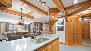 A spacious kitchen with wooden cabinets, a large island with a sink, and a chandelier. There is a stone wall and a view of the outdoors through large windows.