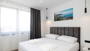 Een moderne hotelkamer met een groot bed, witte lakens, een schilderij van een kustlandschap en een nachtkastje met een afstandsbediening.