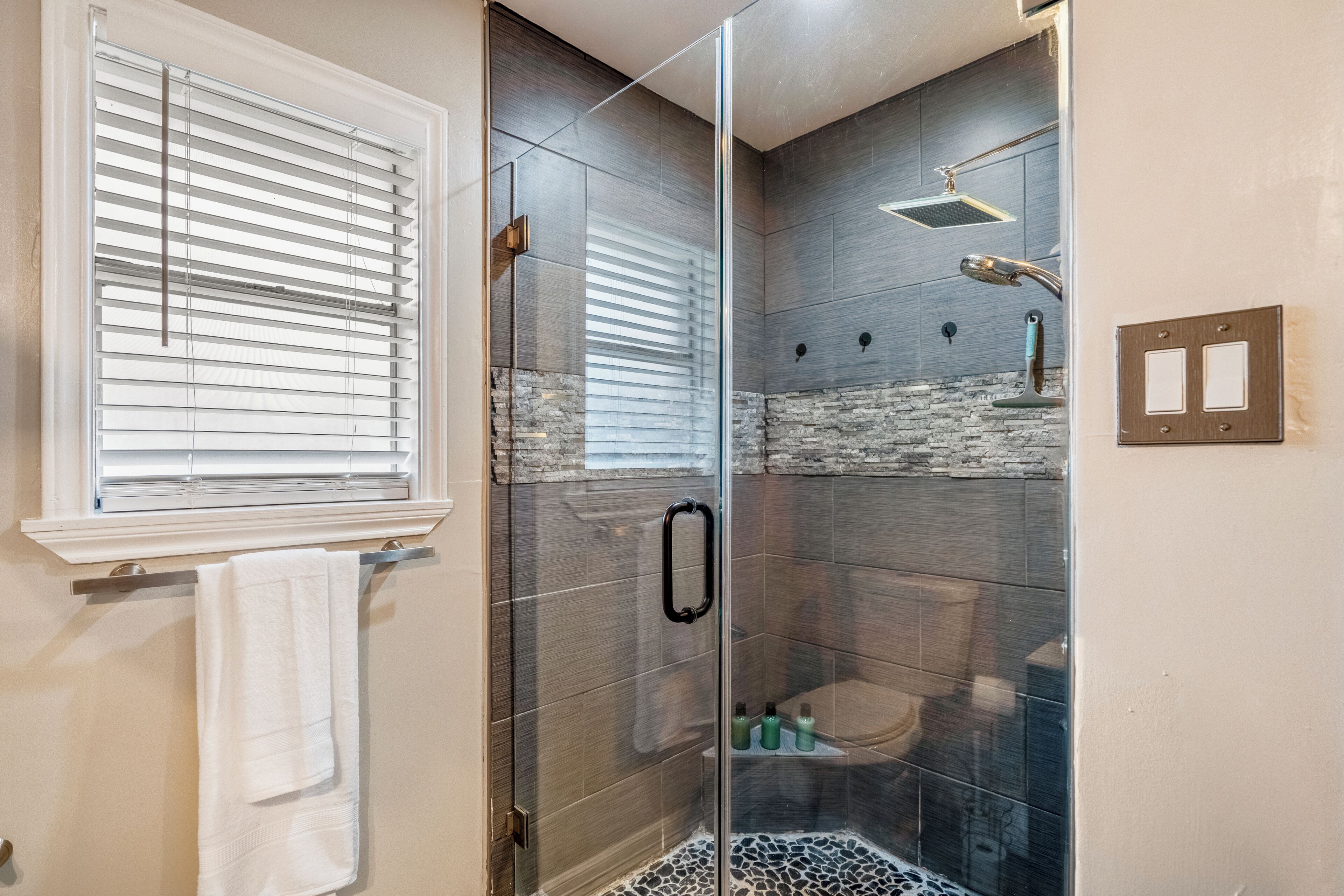 Un baño moderno con ducha envuelta en vidrio, una toalla blanca colgada en un perchero y una ventana con persianas.