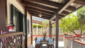 Un patio couvert doté d’un sol en pierre, de meubles en bois et offrant une vue sur une clôture blanche et une végétation luxuriante en arrière-plan.