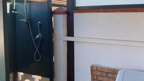 Un espace de douche extérieur abrité, équipé d’un tuyau et d’un pommeau de douche en métal, d’un mur en briques et d’une porte blanche.