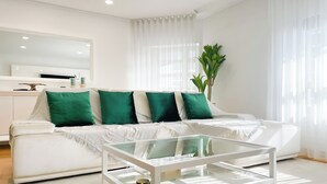 A modern living room with a white sofa, green cushions, a glass coffee table, and a large window with sheer curtains. - Prestige Lisbon Entire 3 Bedroom Luxury Family Friendly Apartment (Lisboa)