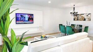 A modern living room with a white sofa, a coffee table, a TV mounted on the wall, a dining area with green chairs, and a chandelier. - Prestige Lisbon Entire 3 Bedroom Luxury Family Friendly Apartment (Lisboa)