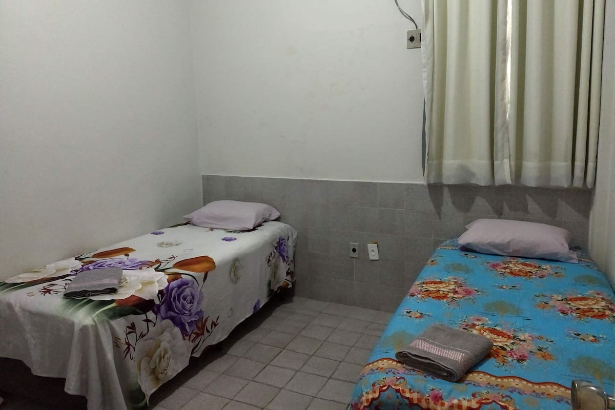Une petite chambre simple, avec deux lits simples, chacun recouvert d’une couvre-lit à motifs floraux. Un climatiseur mural est fixé au-dessus des lits, et une rideau est accroché à la fenêtre.