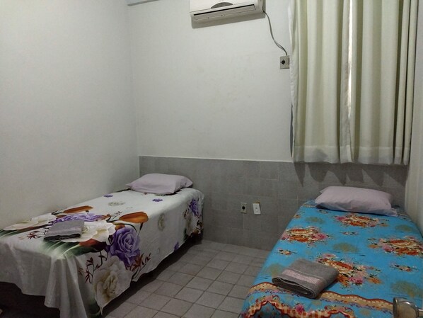 Una habitación pequeña y sencilla con dos camas individuales, cada una con colcha estampada. Hay un aire acondicionado de pared sobre las camas y una cortina en la ventana.