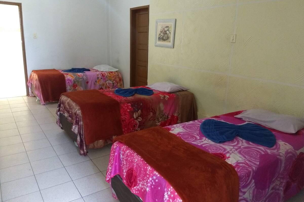 Une chambre avec trois lits, chacun recouvert d’une courtepointe à motifs floraux et d’une couverture marron. Il y a une porte sur la droite et un mur orné d’un petit tableau encadré.