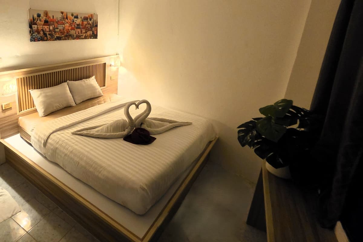 A neatly made bed with a heart-shaped towel and two pillows, a bedside plant, and a wall art piece above the bed.