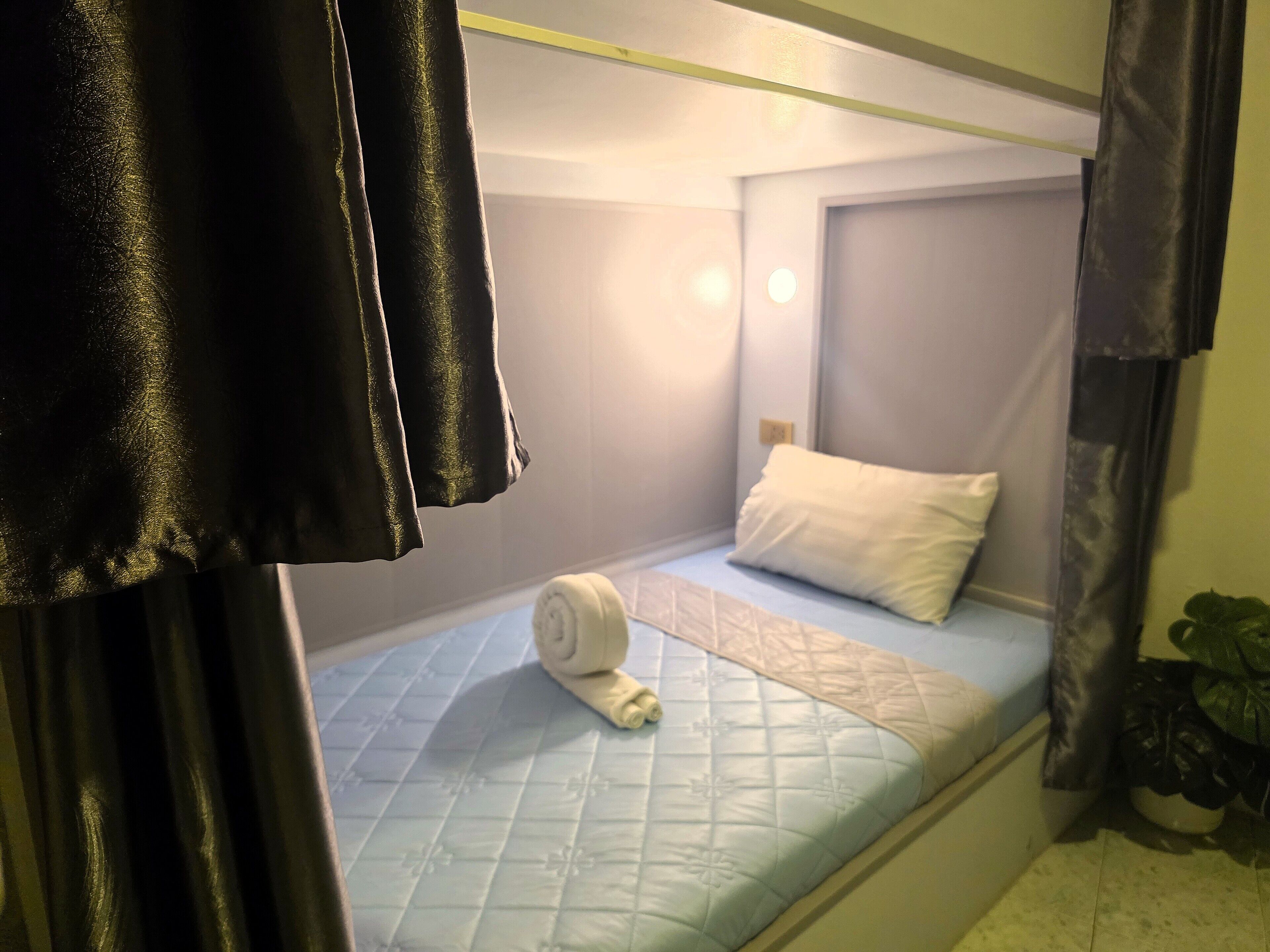 A small, single-bed room with a quilted mattress, a rolled towel, and a white pillow.