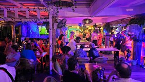 A crowded indoor venue with tables and chairs, a stage with performers, and a neon sign. - Hideout Hostel 2 (Ko Pha-ngan)