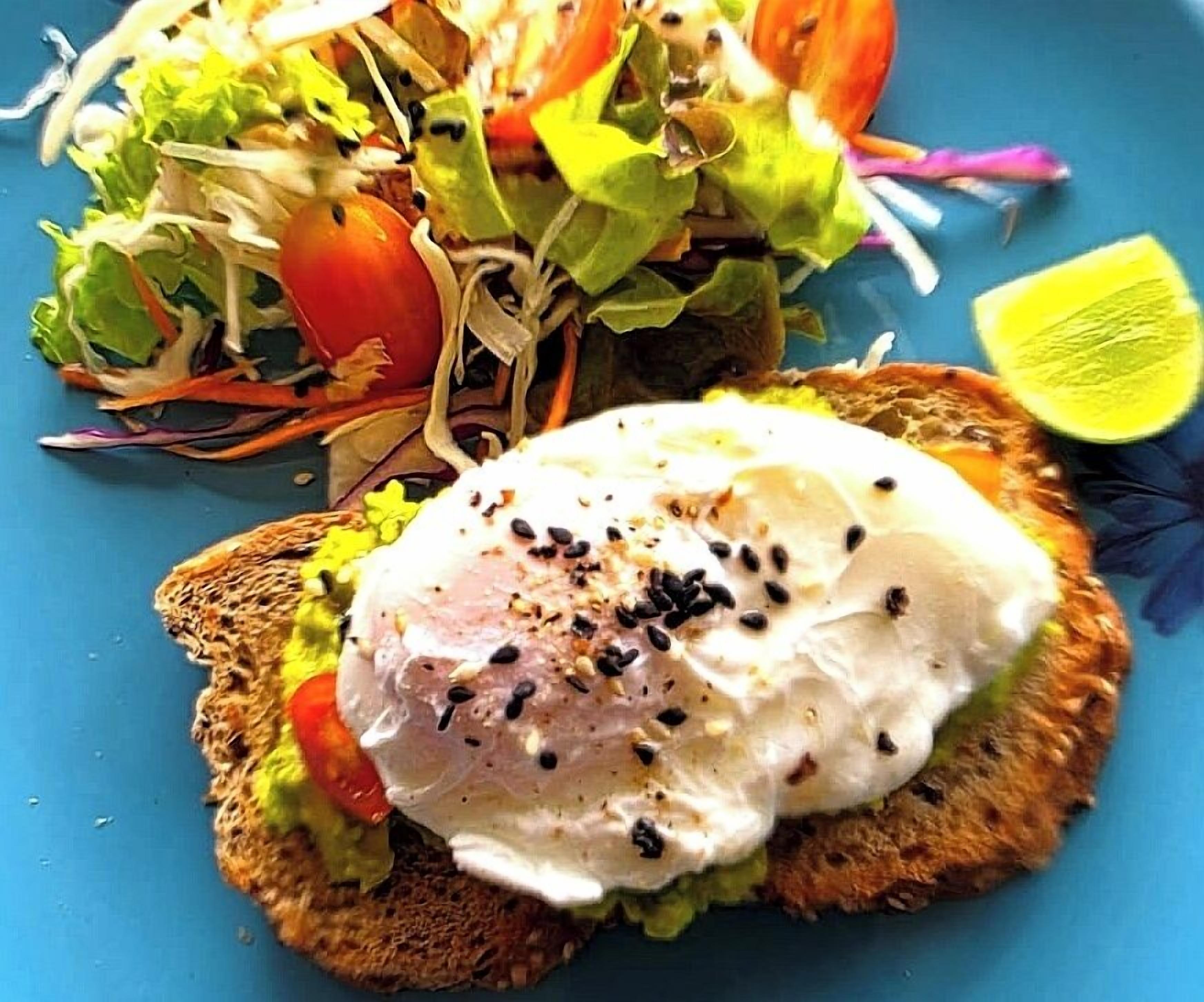 Un sándwich con un huevo escalfado sobre tostada, una ensalada pequeña y una rodaja de lima en un plato azul.