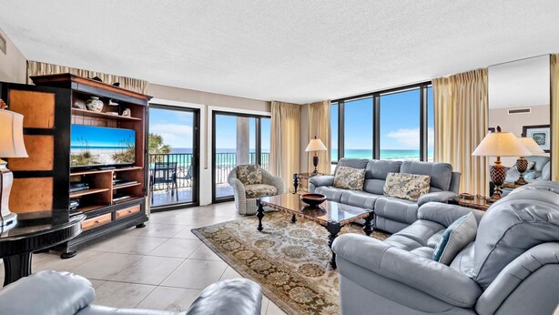 A spacious living room with a large sofa set, a coffee table, and a TV. There is a bookshelf with decorative items and a view of the ocean through large windows.