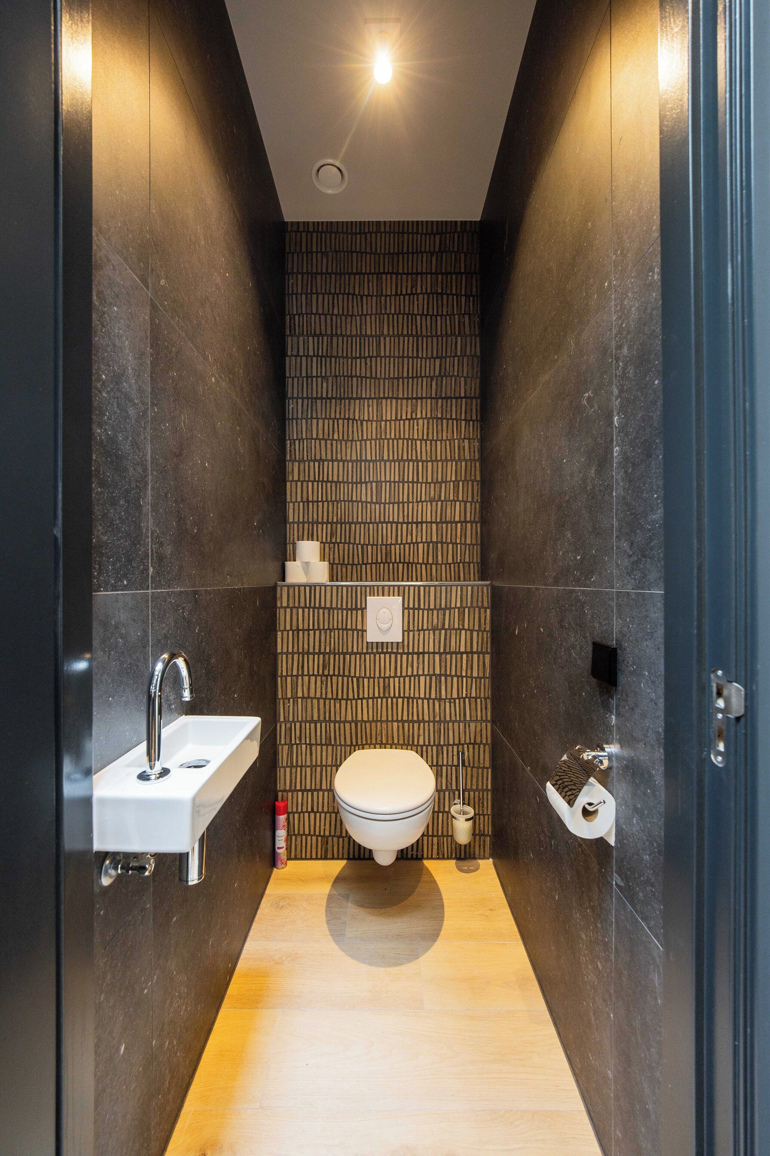 A narrow, modern bathroom with a wooden floor, a white sink, and a toilet.