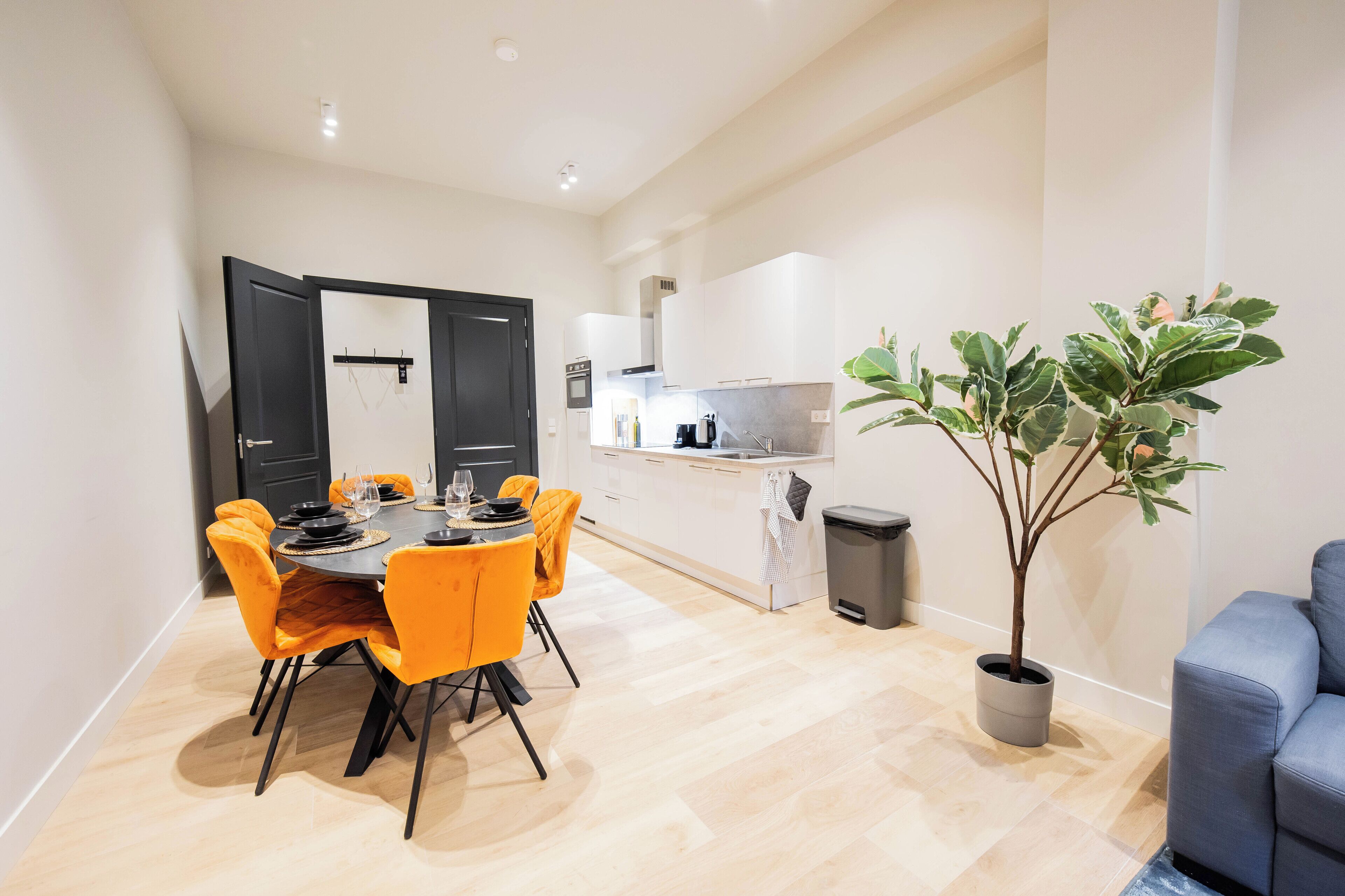 A modern kitchen with white cabinets, a dining area with orange chairs, and a potted plant.