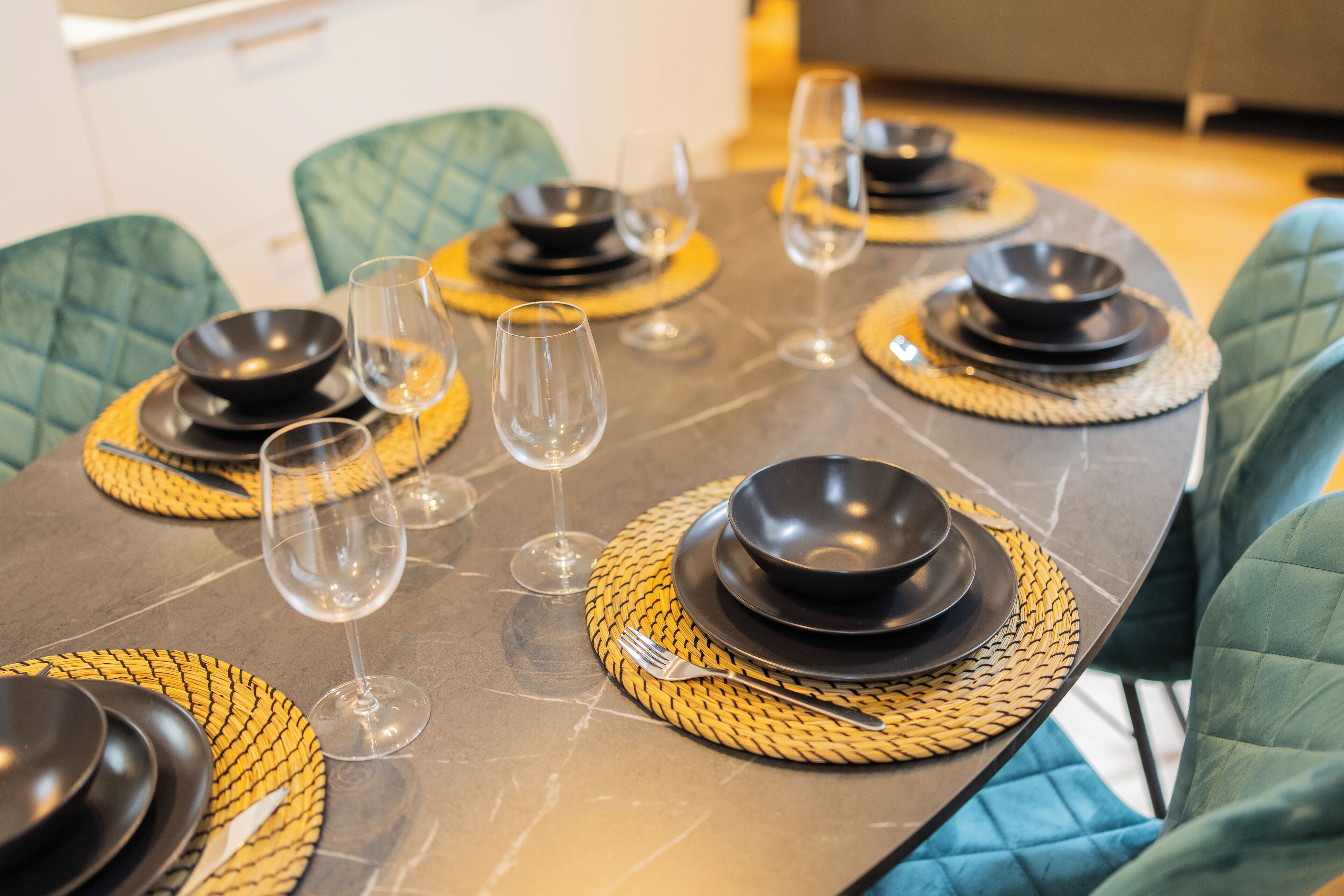 Mesa de comedor dispuesta con platos, cuencos y copas de vino negros, rodeada de sillas tapizadas en verde.