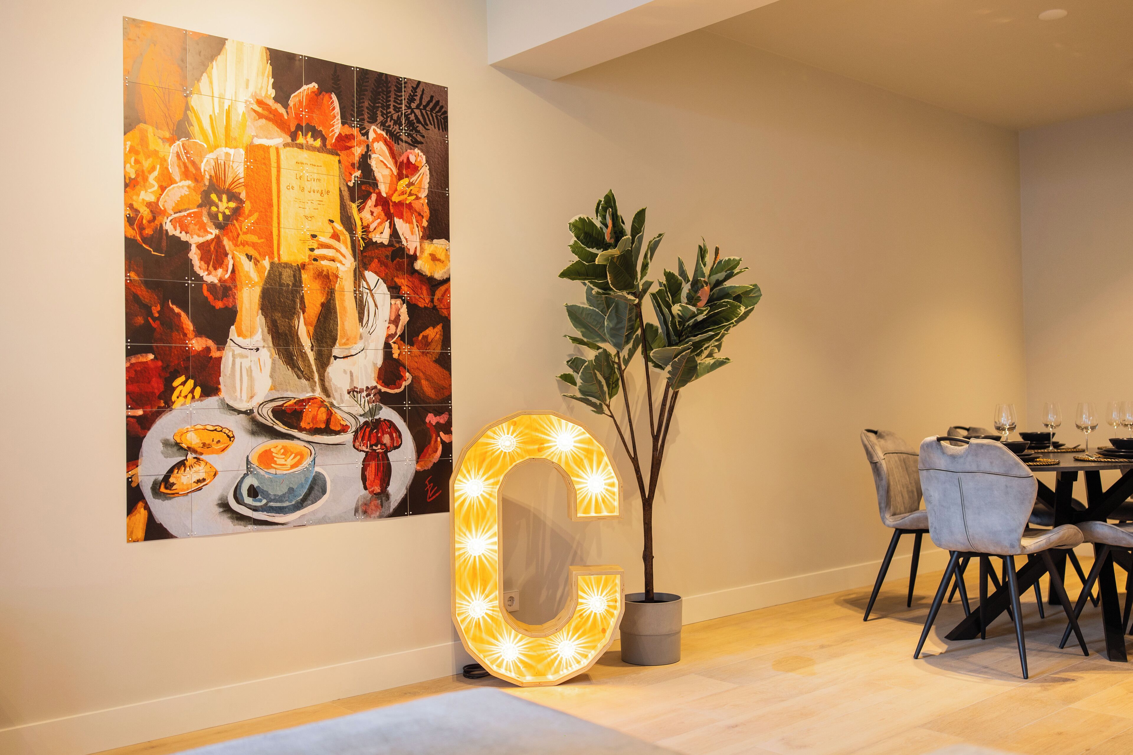 Een eetgedeelte met een grote, verlichte letter 'G', een potplant en een levendig schilderij aan de muur.