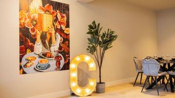 Een eetgedeelte met een grote, verlichte letter 'G', een potplant en een levendig schilderij aan de muur.
