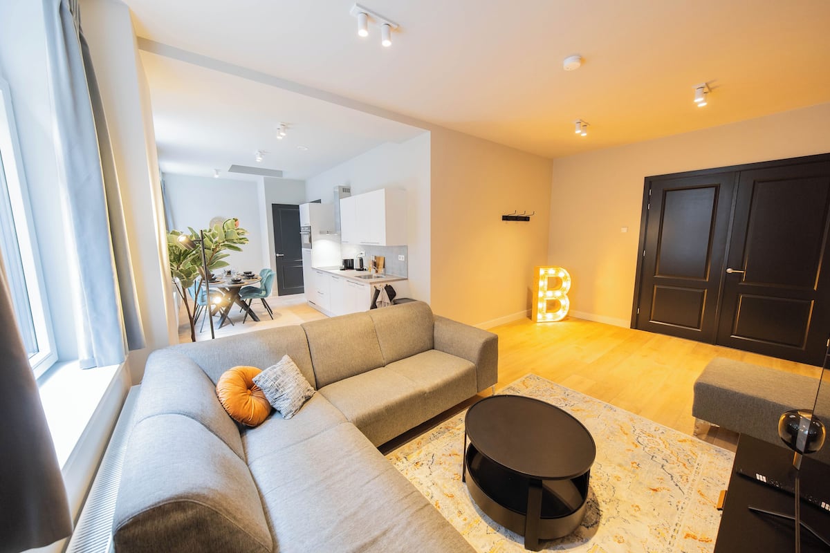 Ein modernes Wohnzimmer mit einem grauen Sofa, einem runden Couchtisch und einem Essbereich im Hintergrund.