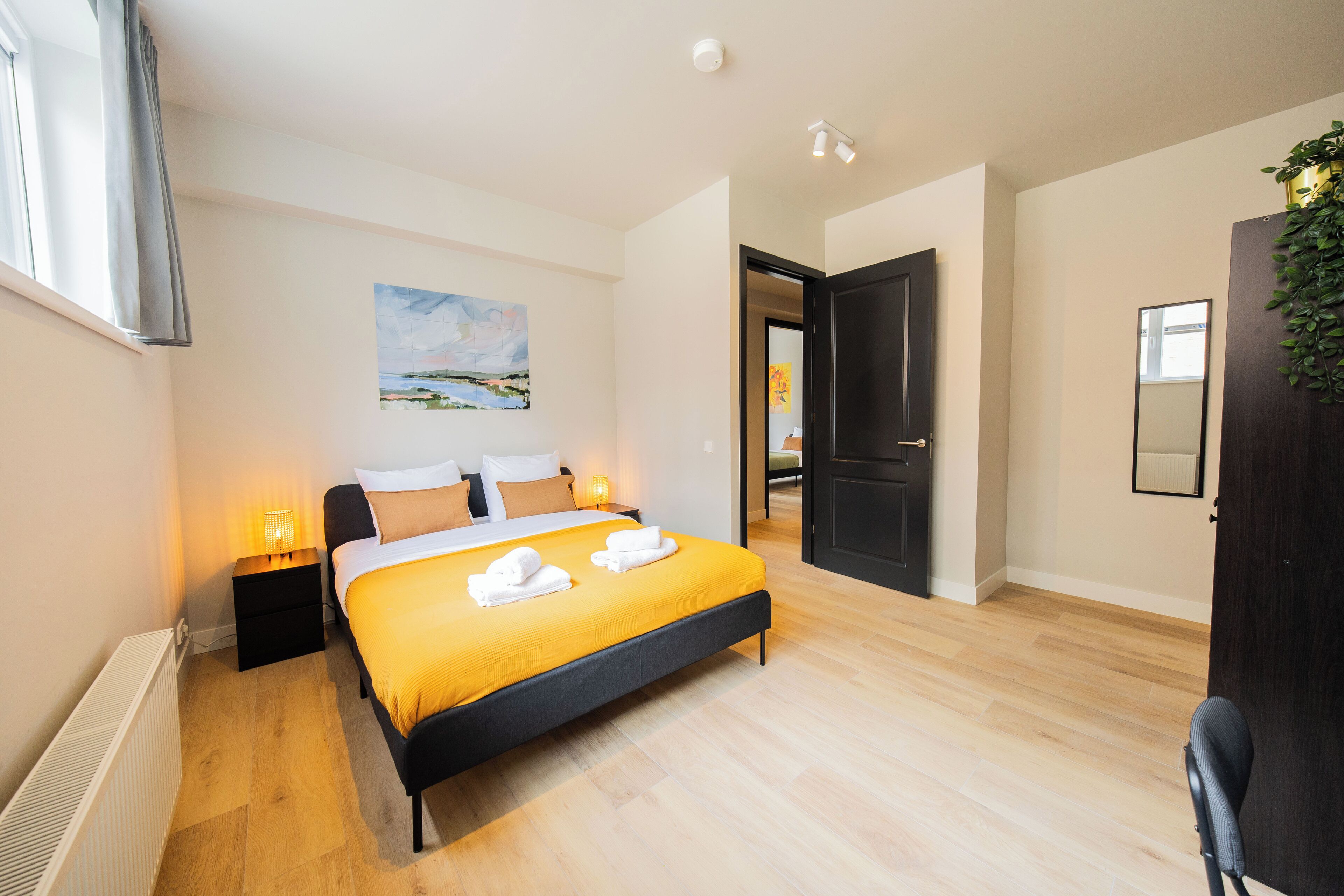 A modern hotel room with a bed, bedside tables, a radiator, a painting, and a door leading to another room.