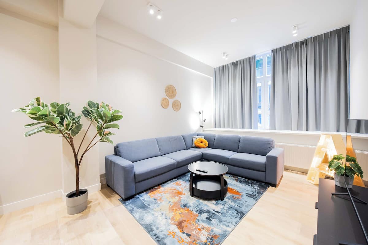 Ein modernes Wohnzimmer mit einer grauen Eckcouch, einem Couchtisch und einem großen Fenster mit grauen Vorhängen. Zwei Topfpflanzen und ein Wanddekor ergänzen das Bild.