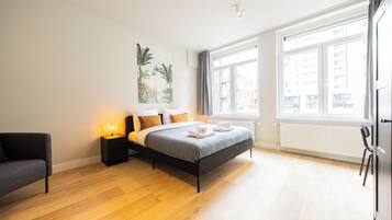 A modern bedroom with a bed, bedside tables, a chair, and large windows.