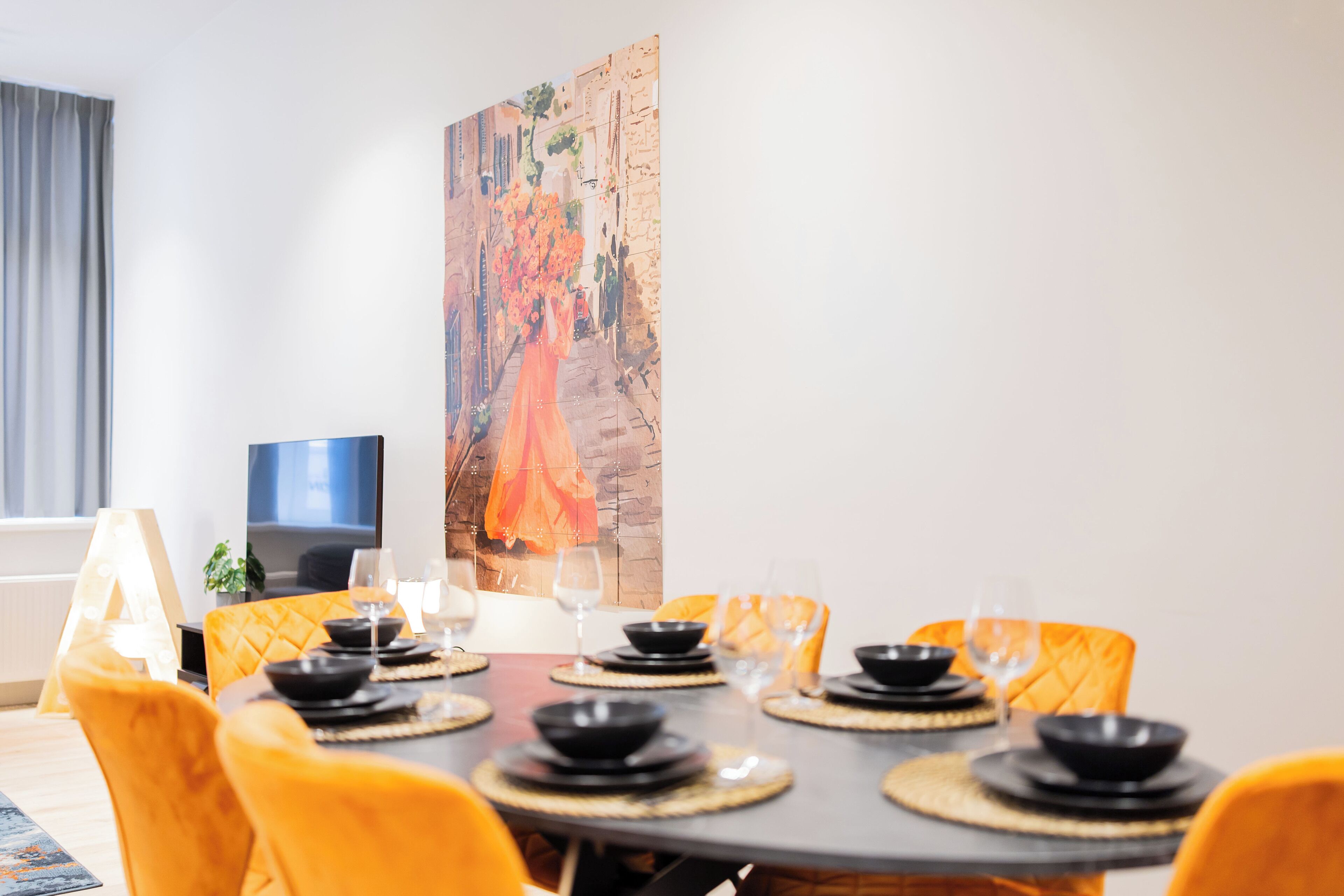 Un coin repas avec une table ronde dressée pour quatre personnes, entourée de chaises jaunes. Un tableau est accroché au mur derrière la table.