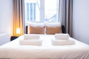 Una cama bien hecha con dos almohadas y dos toallas dobladas, una ventana con cortinas y una lámpara sobre una mesita de noche.