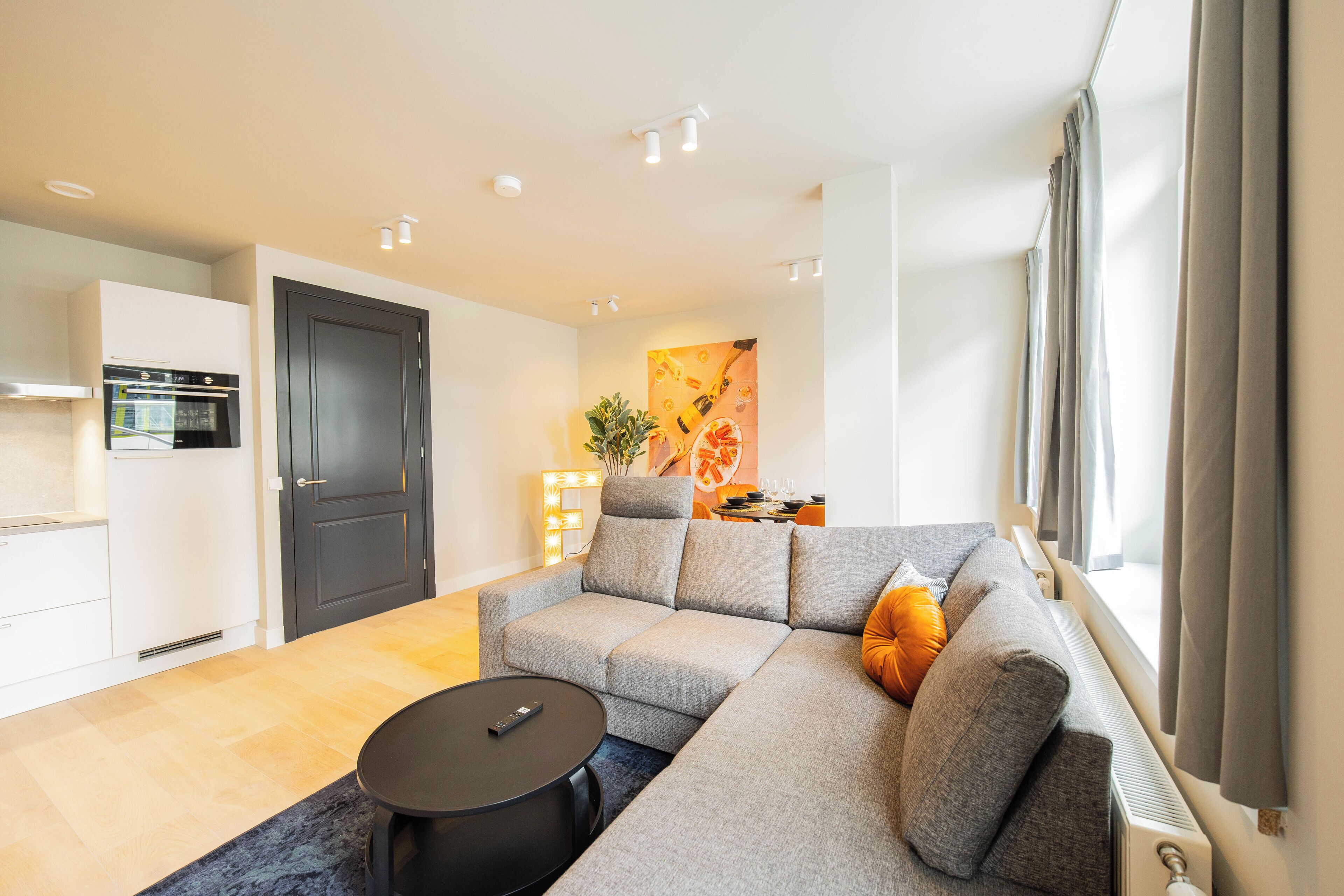 Een moderne woonkamer met een grijze hoekbank, een klein zwart tafeltje en een keuken met een ingebouwde oven en magnetron.