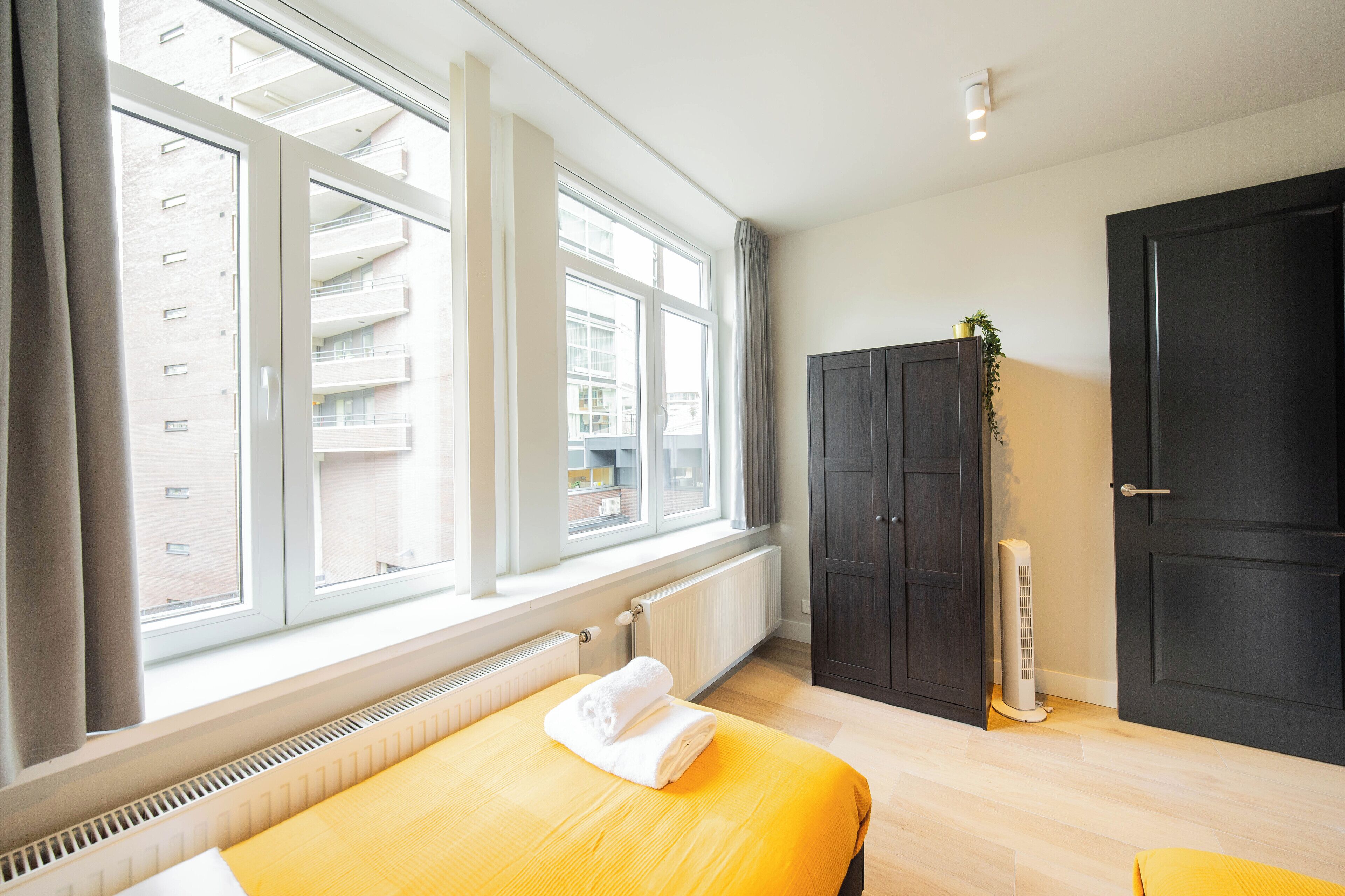 Een hotelkamer met een bed, een radiator, een raam met gordijnen, een donkere kast en een radiator op de vloer.