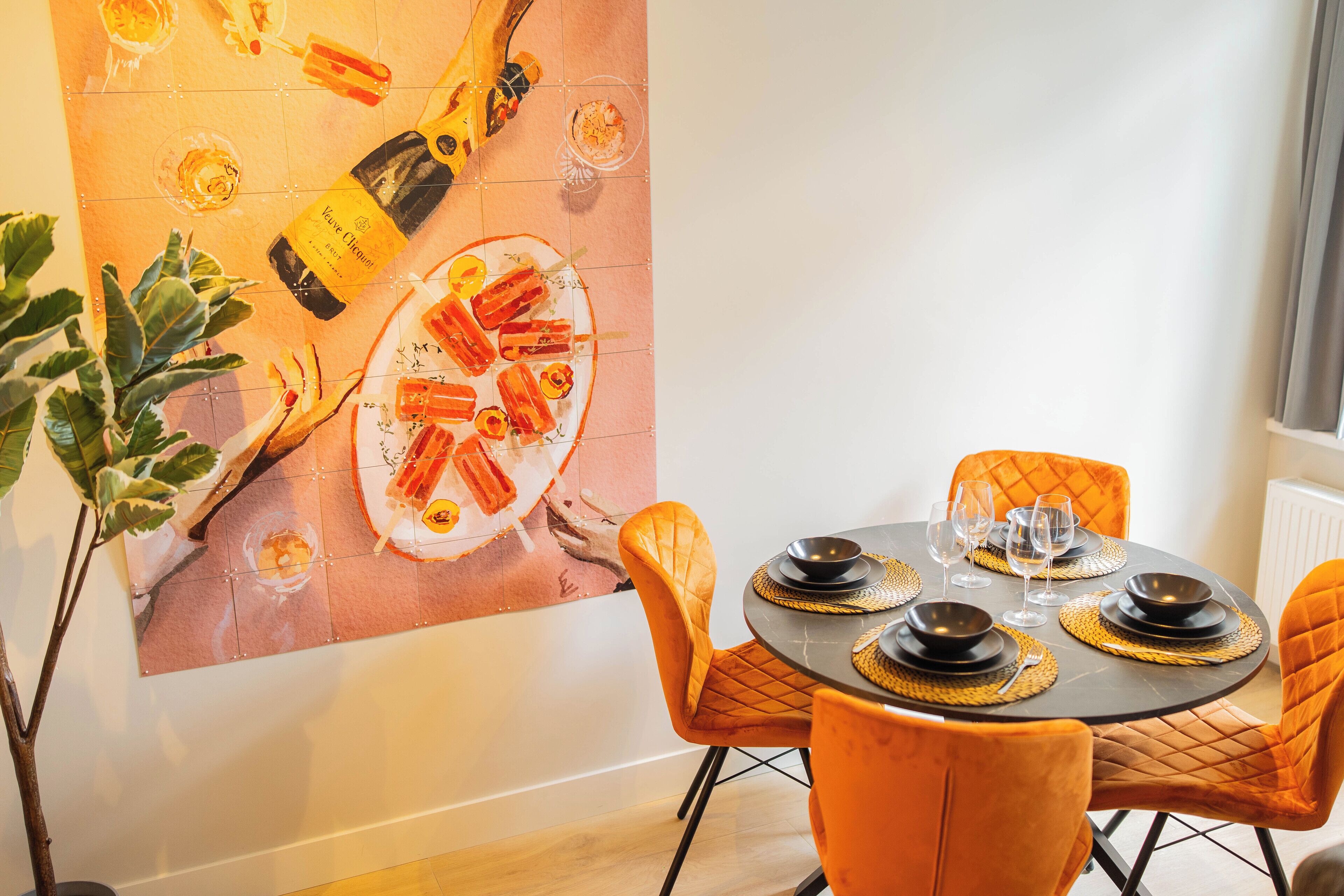 A dining area with a round table set for four, orange chairs, and a large painting on the wall depicting food and drink.