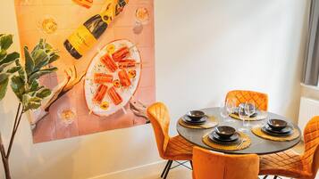 A dining area with a round table set for four, orange chairs, and a large painting on the wall depicting food and drink.