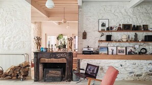 Un salon chaleureux avec une cheminée, une table basse en bois, un fauteuil rouge et un mur en pierre agrémenté d’étagères où sont exposés divers objets.