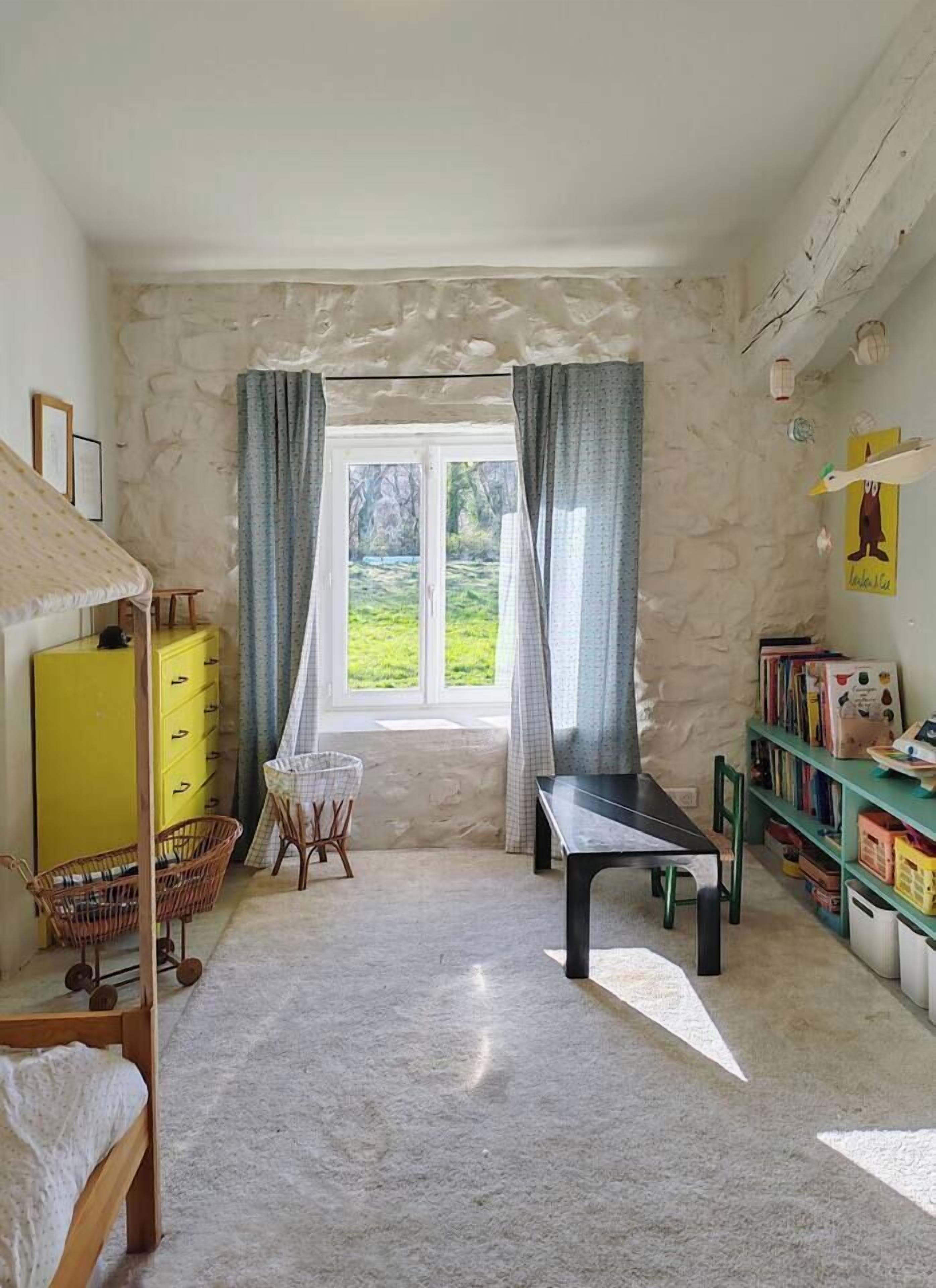 Une pièce comprenant un lit, une table, une chaise, une bibliothèque et une fenêtre avec des rideaux.