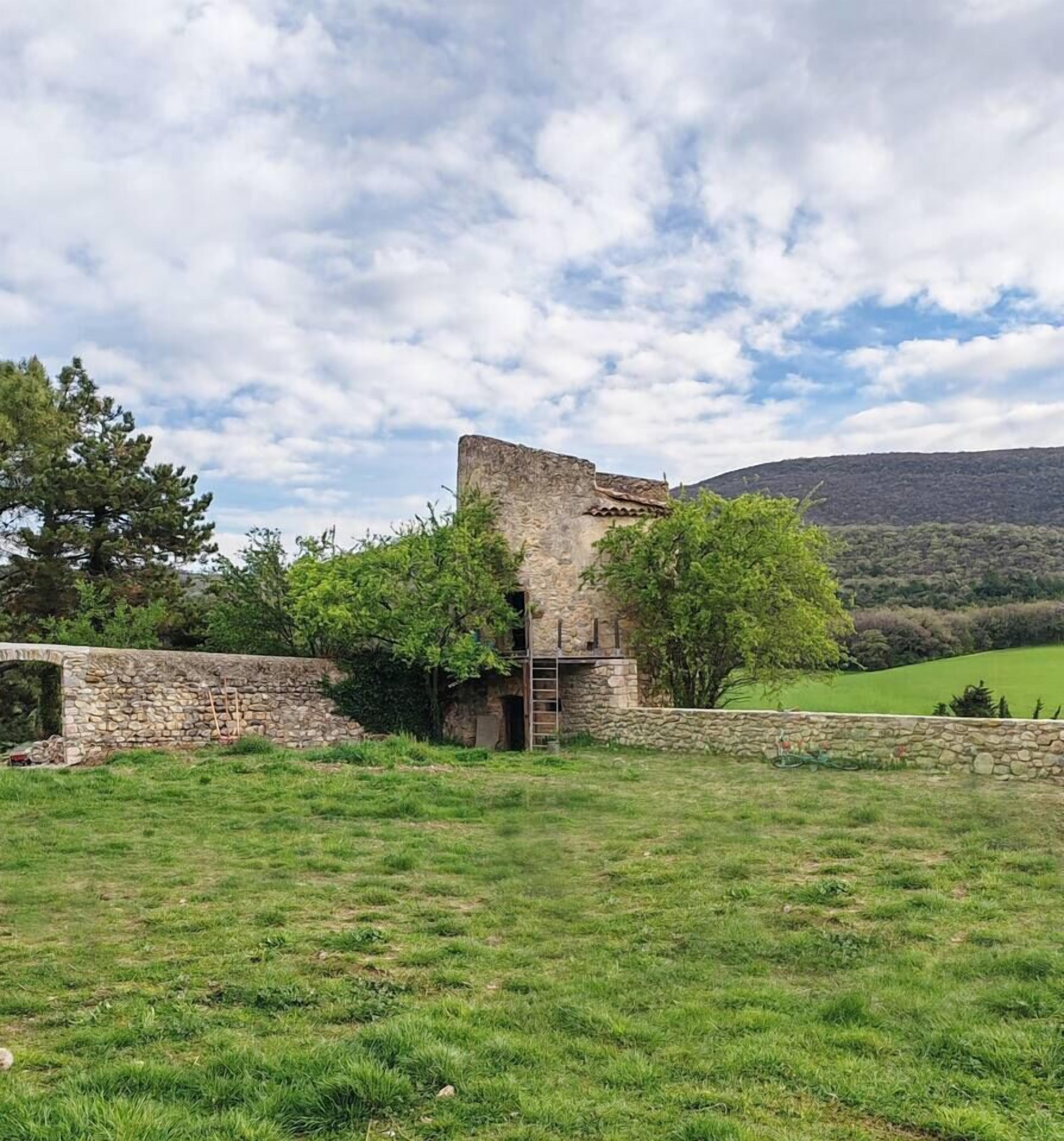 A rustic stone building with a sloped roof, surrounded by a stone wall and lush greenery.