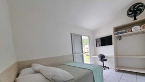 Ein Einzelbett mit Kissen, ein Schreibtisch mit Stuhl, ein an der Wand befestigter Fernseher und ein Ventilator auf einem Regal.