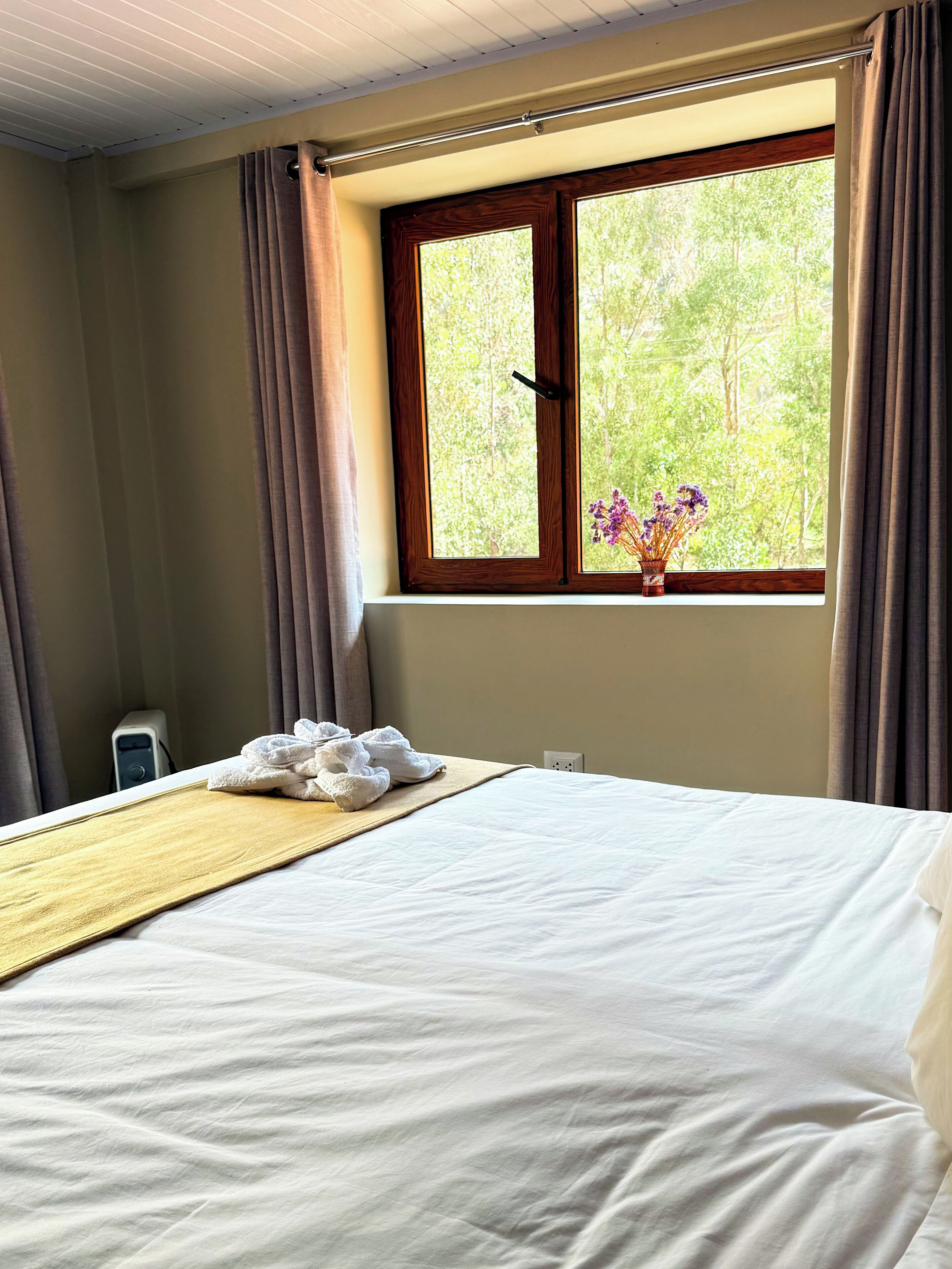 Een net opgemaakt bed met witte lakens, een houten hoofdbord en een raam met uitzicht op groen.