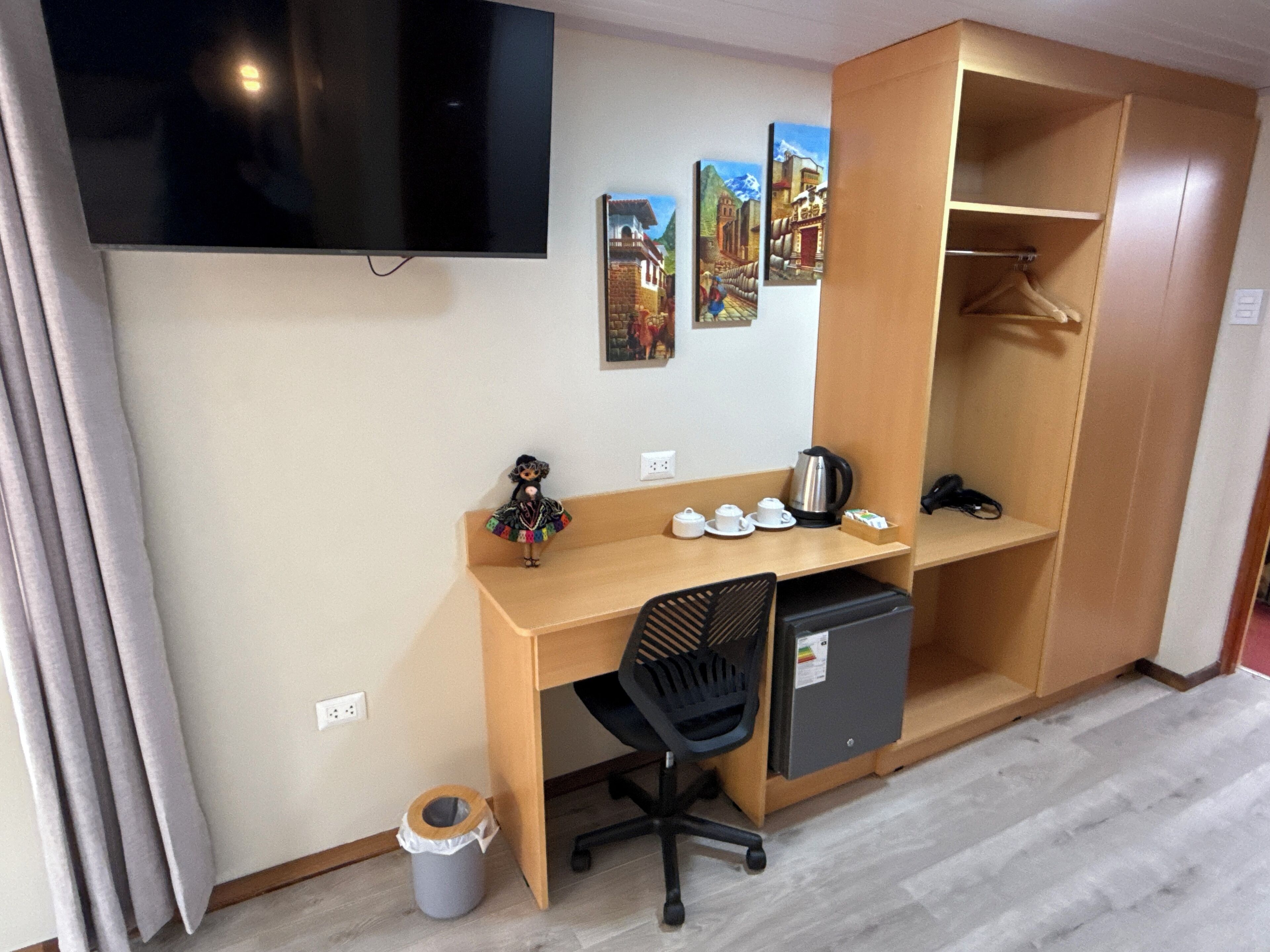 A compact hotel room with a wooden desk, a black office chair, a small refrigerator, a TV mounted on the wall, and a wardrobe with hanging space.