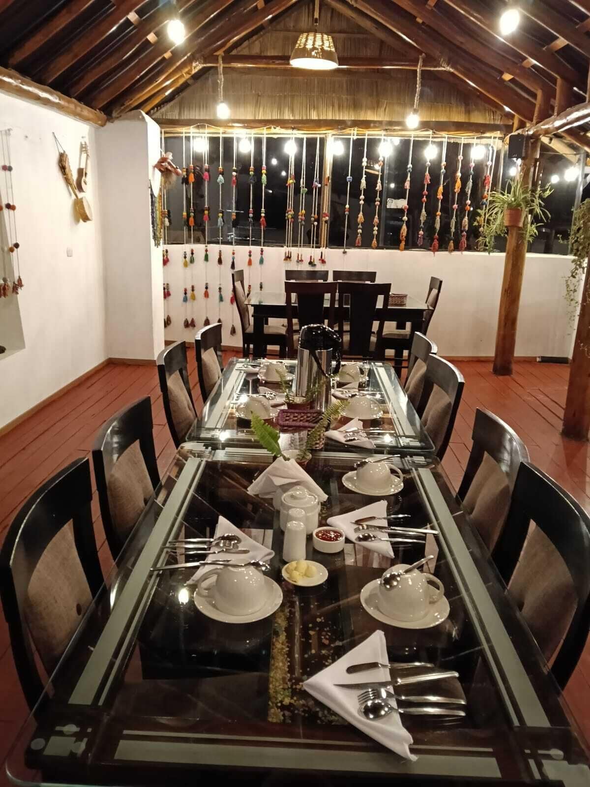 Un comedor con una larga mesa dispuesta para seis personas, rodeada de sillas. La mesa tiene una cubierta de vidrio y diversos platos y utensilios ordenados. La habitación presenta un techo de madera rústica y paredes decoradas con adornos colgantes.